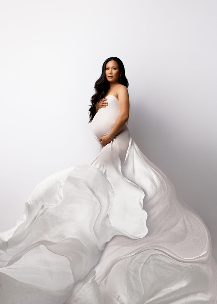 Maternity Photography, pregnant woman in long white flowing dress against white backdrop